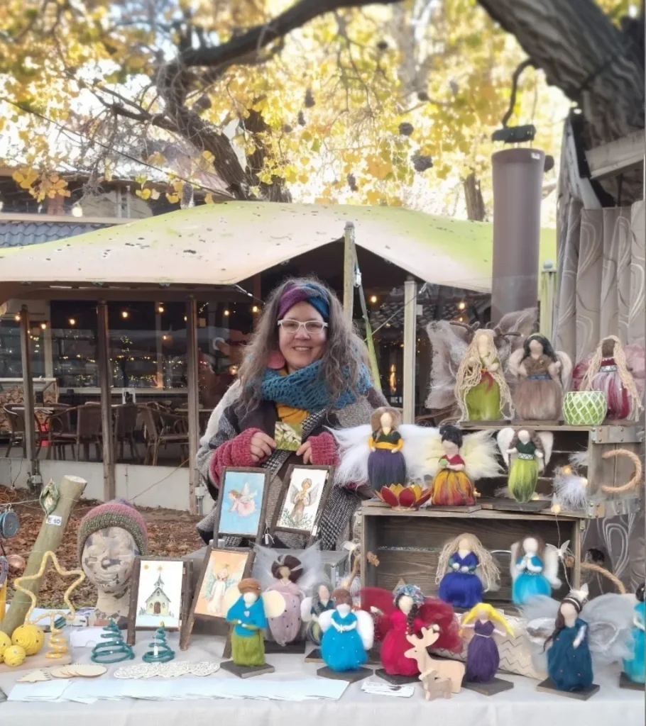 Maria sitting at her handmade craft market booth displaying dozens of colorful needle felted angels and ornaments on wooden shelves, with autumn leaves and string lights creating a warm atmosphere