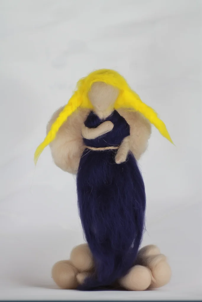 MariaElena | Handmade needle felted angel with flowing golden yellow hair and navy blue dress, holding a small dark companion close to her chest, standing on cream-colored felted stones